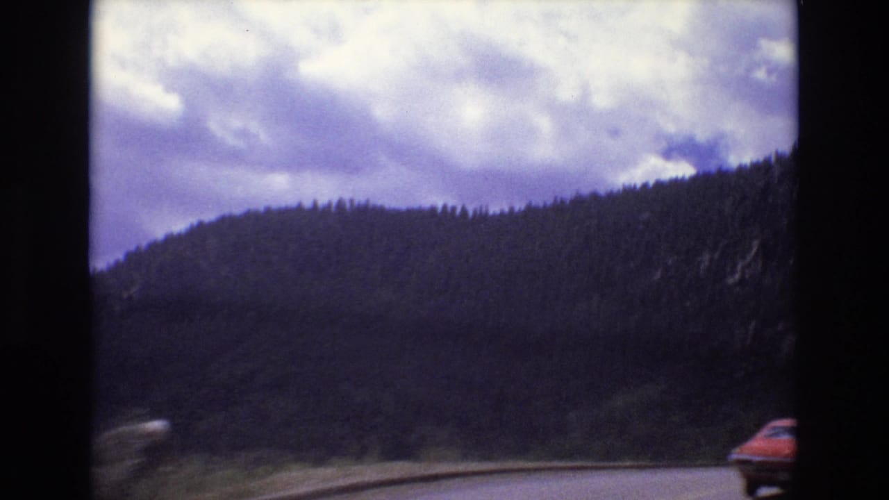Red car partly at frame edge on a road through forested mountains.