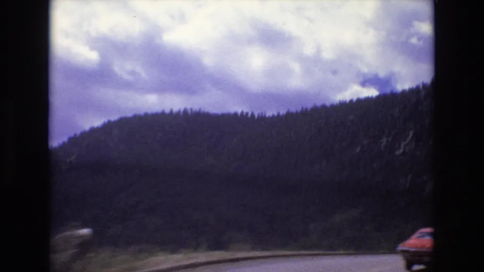 1973 Colorado road-trip frame with a red car near the right edge, a paved mountain road, dark evergreen hills, and cloud cover overhead.