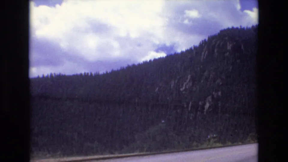 1973 Colorado 8mm view of a forested mountainside, rocky outcrops, and the edge of a winding paved road filmed from a moving car.