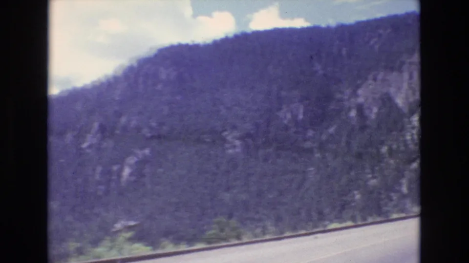 1973 Colorado home-movie frame showing the lower edge of a mountain road, dense tree-covered slopes, rocky patches, and bright clouds.