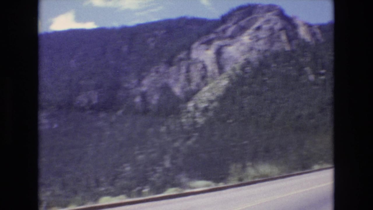 1973 Colorado 8mm frame showing a curving mountain highway below evergreen-covered slopes and exposed rock under a blue sky.