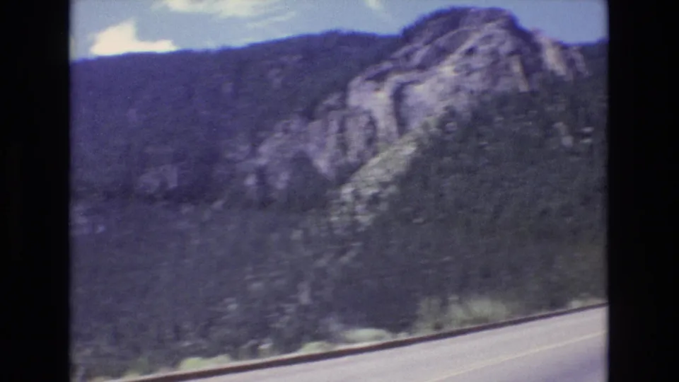1973 Colorado 8mm frame showing a curving mountain highway below evergreen-covered slopes and exposed rock under a blue sky.