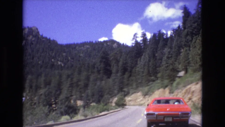 1973 Colorado 8mm frame showing the rear of a red car on a winding highway through dense evergreens and rocky mountain terrain.