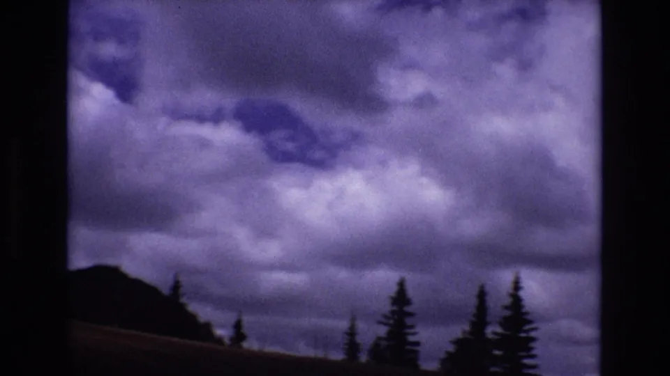 1973 Colorado 8mm frame with dense purple-tinted clouds over a dark ridge and several silhouetted evergreen trees.
