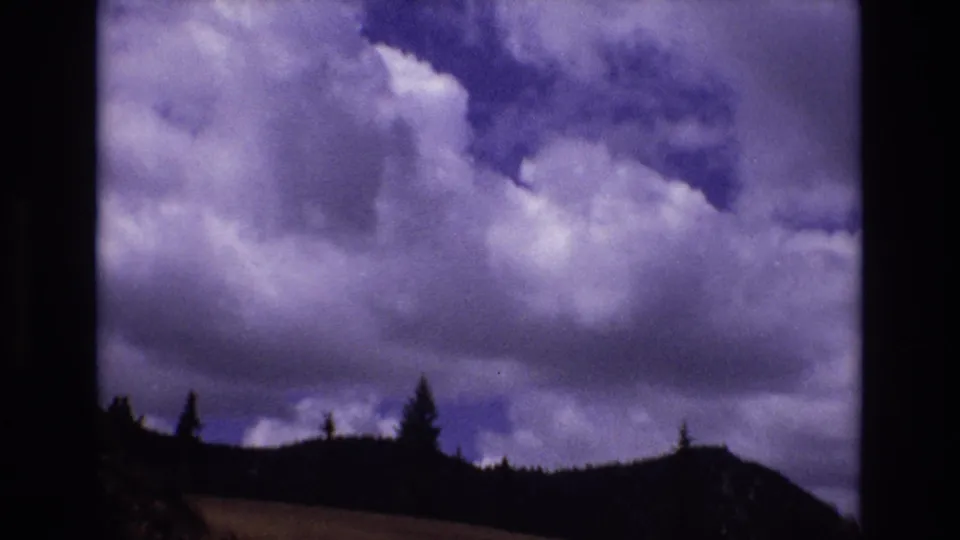 1973 Colorado 8mm frame with dramatic cloud cover hanging over a shadowed ridge and scattered evergreens.