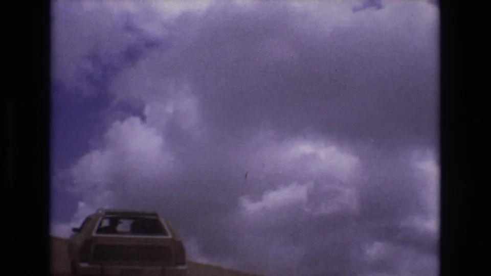 1973 Colorado 8mm frame showing the rear of a boxy car on a hillside road under purple-shifted clouds.