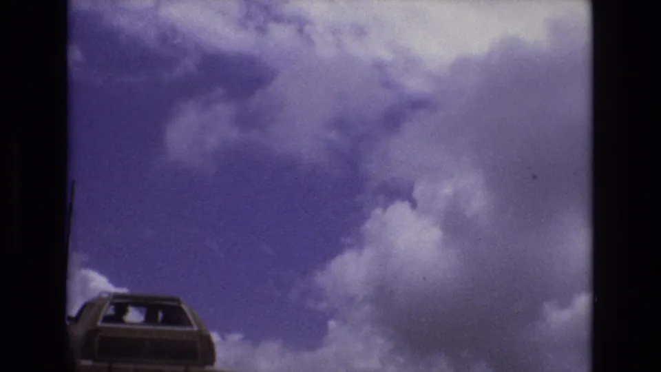 A station wagon appears near the bottom edge of a 1973 Colorado 8mm frame under a broad purple-blue sky filled with large white clouds.
