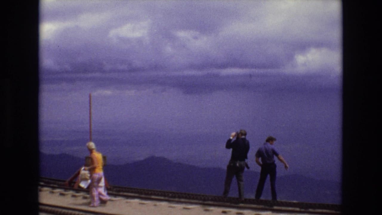Adults standing and walking on a rail overlook with mountains and heavy clouds beyond.