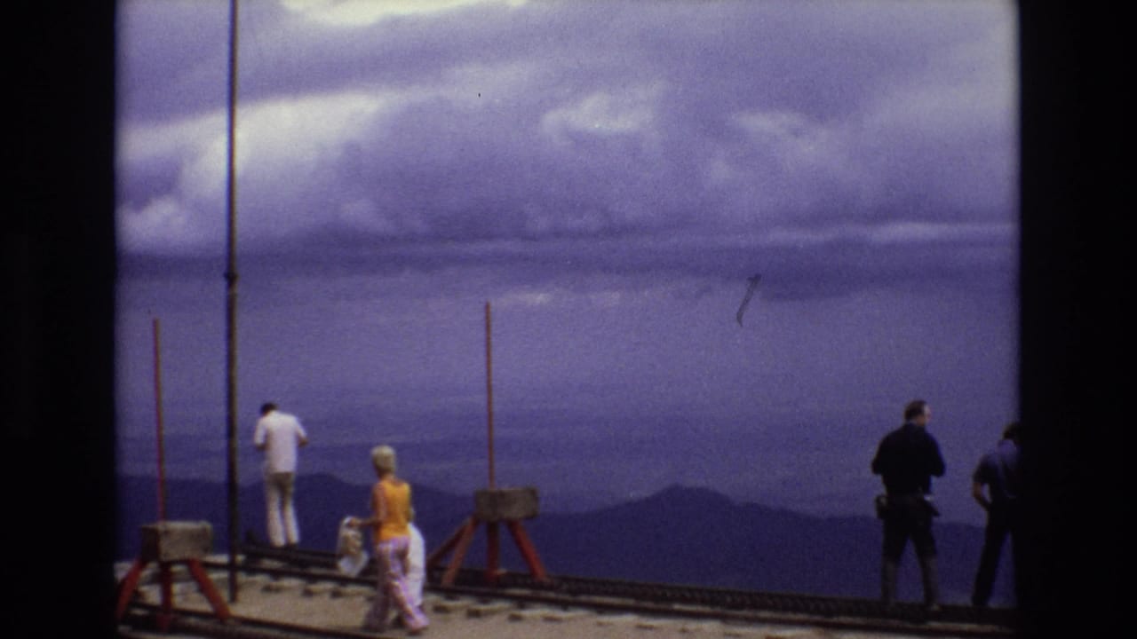 People spaced along a rail platform looking toward a hazy mountain horizon.