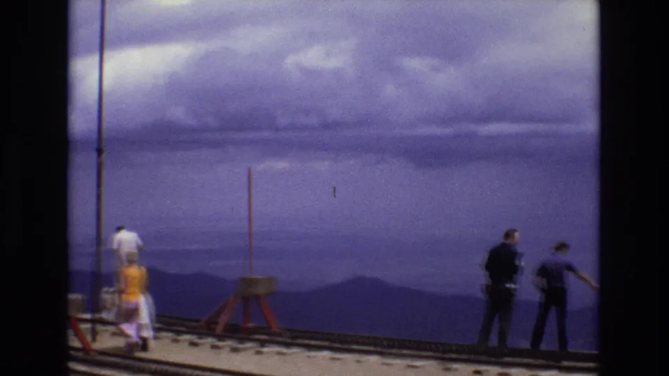 1973 Colorado 8mm frame showing adults standing and walking on a rail overlook, with vertical poles, track rails, and layered mountain silhouettes in the background.
