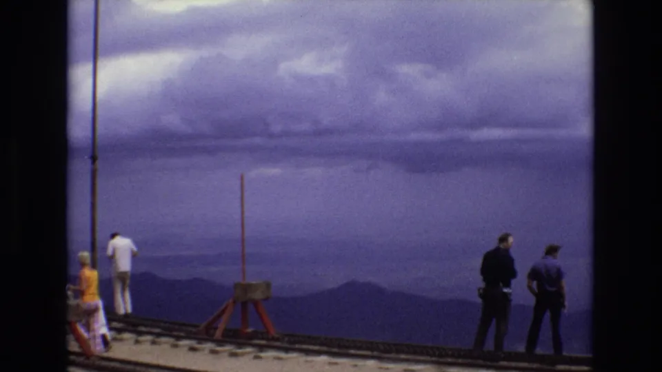1973 Colorado home-movie frame with adults positioned along an elevated rail or bridge area, looking across hazy ridges beneath a purple-tinted overcast sky.