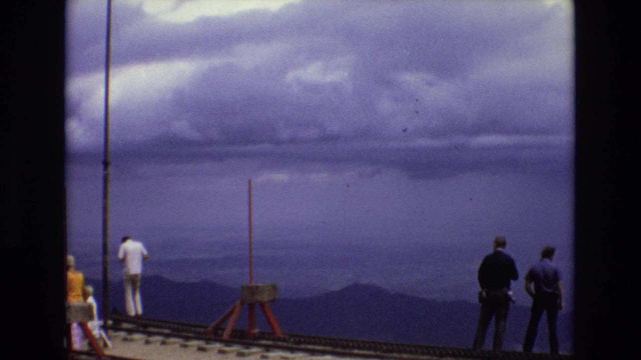 1973 Colorado home-movie frame showing several visitors on an overlook deck facing distant mountain ridges beneath a heavy purple-blue cloud layer.