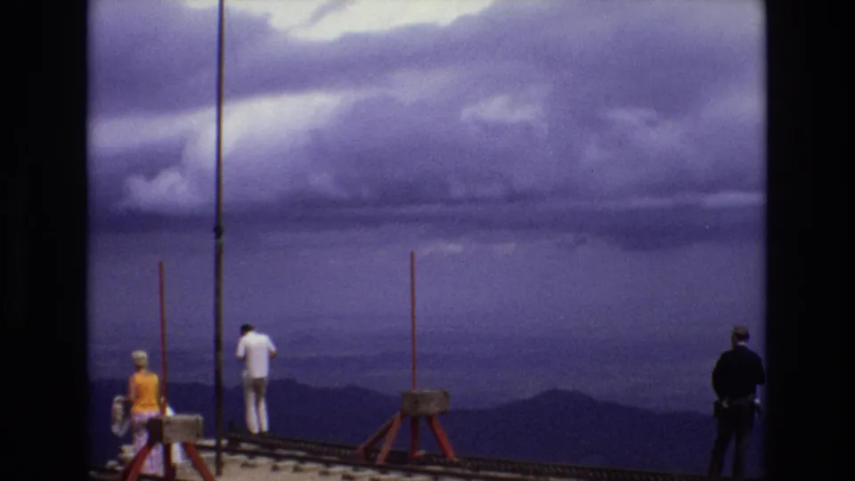 1973 Colorado frame with a man in white, a person in yellow, and another visitor standing near red posts on a scenic overlook above layered mountains.