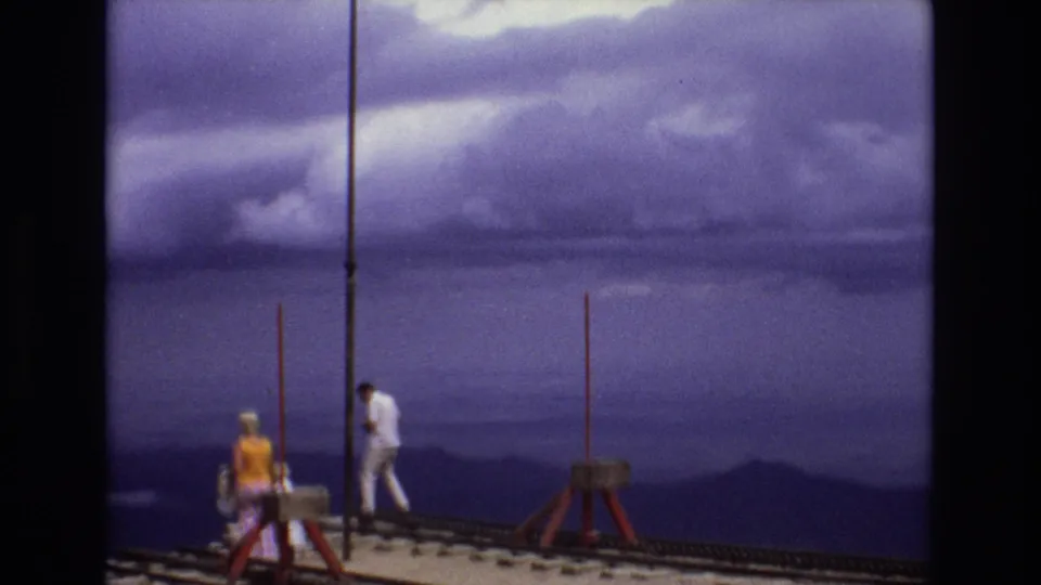 1973 Colorado home-movie frame showing a man in white moving along the overlook as other visitors remain near the edge with cloudy mountains behind them.