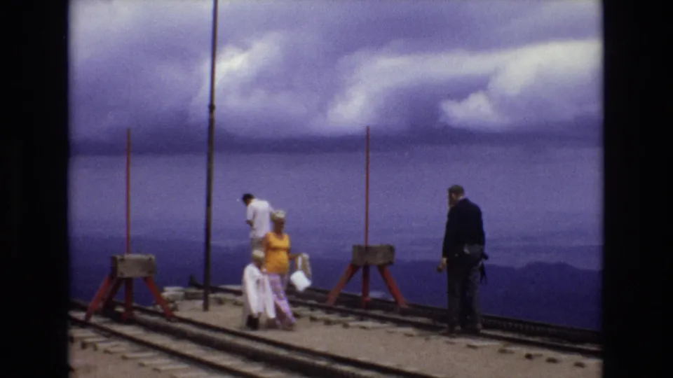 1973 Colorado frame showing visitors clustered on an overlook deck, including a person in yellow seated or leaning near the center and a dark-clothed adult at right.