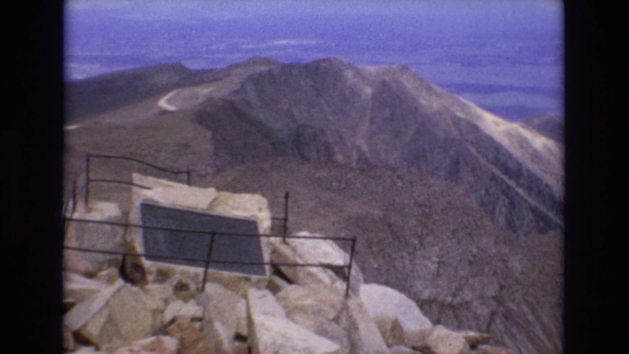 1973 Colorado 8mm frame showing a rocky summit overlook with a metal railing and dark plaque in front of barren ridges and a winding mountain road.