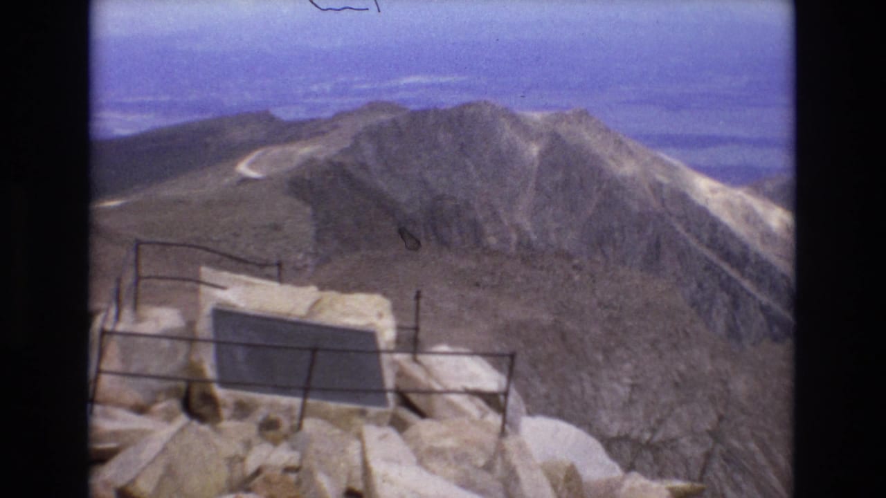 Rocky overlook and railing facing a rugged mountain ridge with a pale winding road beyond.