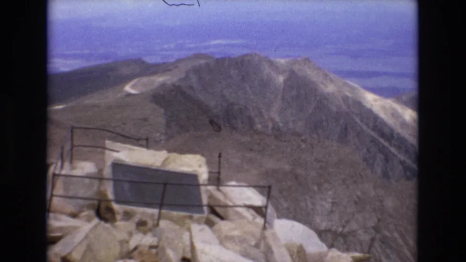 1973 Colorado 8mm frame from a high rocky viewpoint, with a railing and plaque in the foreground and a pale road curving across the distant ridge.