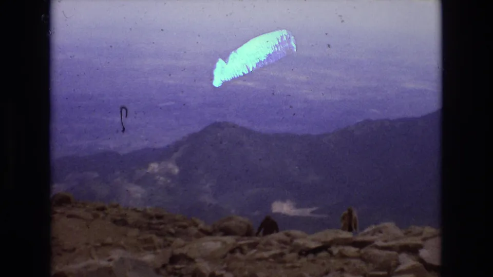 1973 Colorado 8mm frame looking across a hazy mountain valley from a rocky summit, with small distant people near the edge and a bright blue-white film or optical streak overhead.