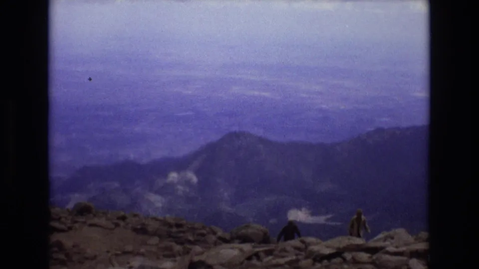 1973 Colorado 8mm frame showing small distant visitors on a rocky summit foreground, looking out across a hazy blue-purple mountain valley and ridge line.