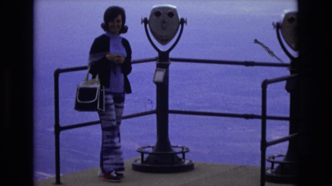 In 1973 Colorado home-movie footage, a woman in a dark cardigan and patterned bell-bottoms stands beside a coin-operated scenic viewer near metal railings.