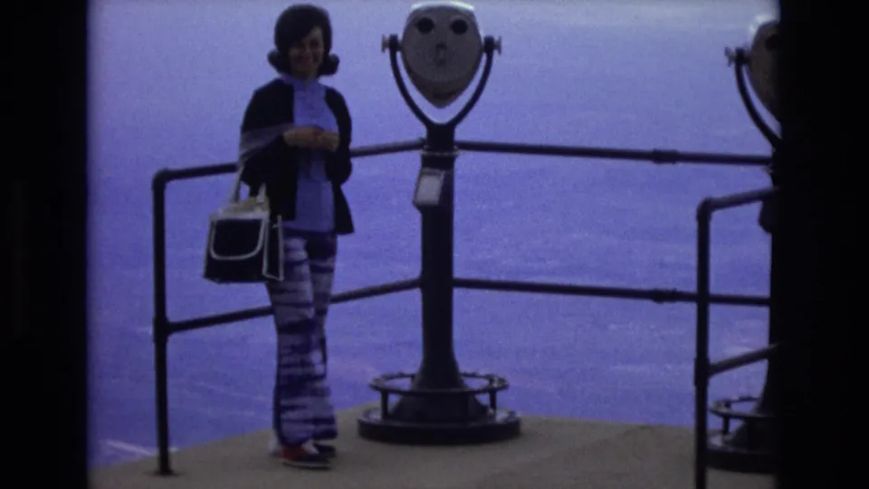 A woman stands near a coin-operated viewing scope and railings at a Colorado scenic overlook, wearing 1970s-style patterned pants and carrying a handbag.
