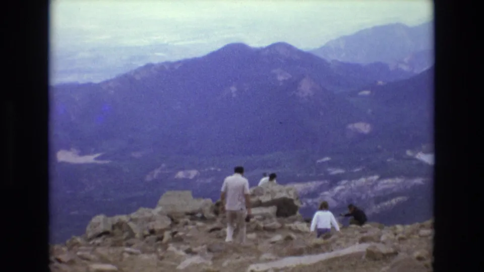 Tourists stand and sit on a rocky Colorado summit or overlook while looking across a hazy mountain panorama in 1973 8mm home-movie footage.