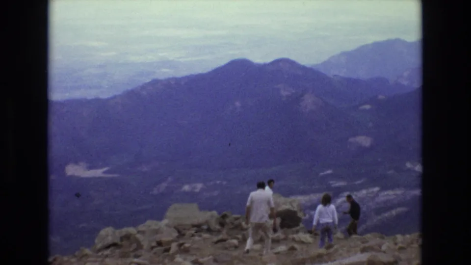 A small group of visitors stands on rocky terrain at a Colorado mountain viewpoint with layered peaks and haze stretching across the background.