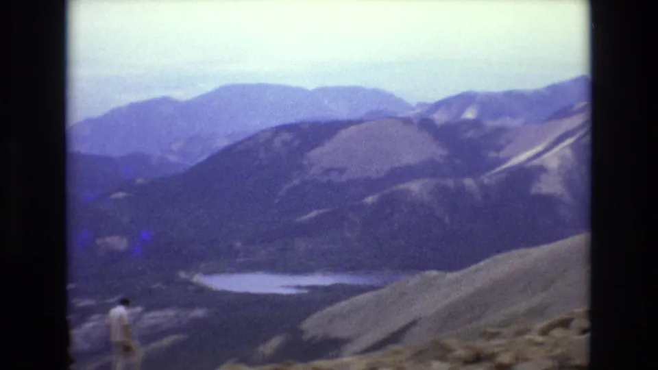 1973 Colorado 8mm frame showing a lone man in light clothing on a rocky alpine slope with mountains and a pale lake or snow patch below.