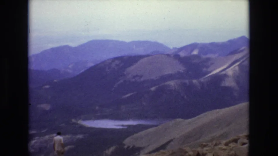 1973 Colorado home-movie frame with a distant man on a rocky ridge facing layered mountains and a pale alpine lake or snow patch.