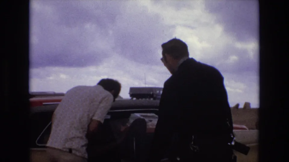 Colorado 1973 8mm frame with two adult men near a classic car, one leaning into the vehicle while another stands in a dark suit under storm clouds.