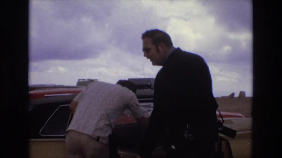 Colorado 1973 roadside home-movie frame showing an adult man in sunglasses and a dark suit beside another man leaning into a car beneath a bright, unsettled sky.
