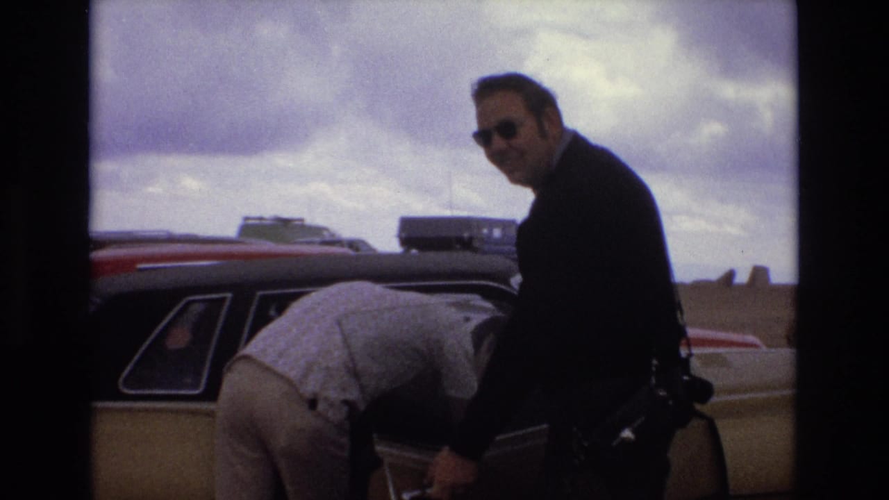 Two adult men beside a car with an open door under a cloudy sky.
