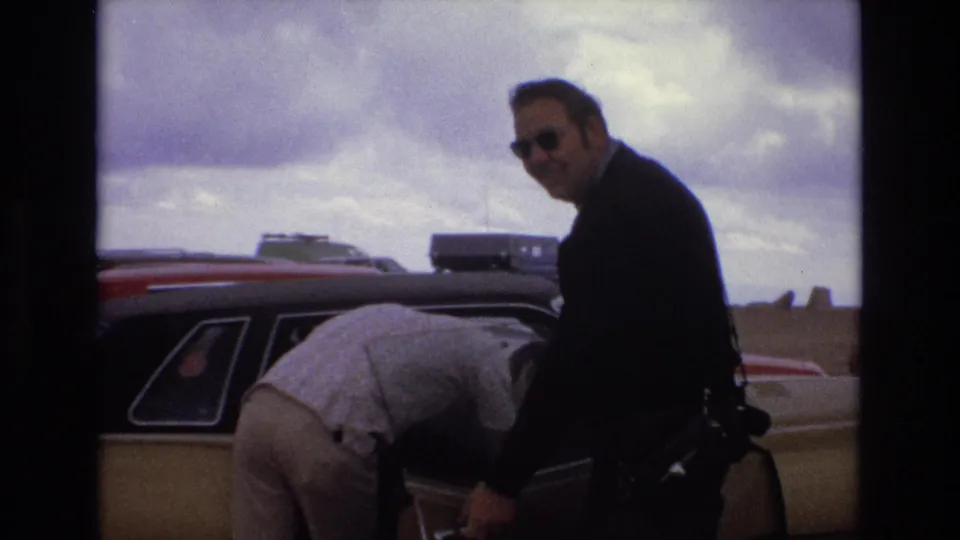 1973 Colorado home-movie frame showing two adult men beside a classic American car, one wearing sunglasses and looking toward the camera while the other bends near the open door.