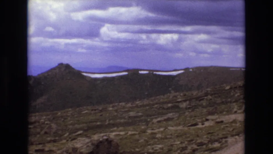 Wide 1973 Colorado mountain view showing a rocky high-country slope, a pointed ridge form, snow patches along the crest, and a broad overcast sky.