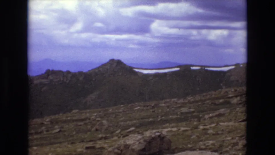 Colorado mountain panorama with a rugged ridgeline, visible snow patches, rocky foreground, and blue-purple 8mm color shift under a cloudy sky.