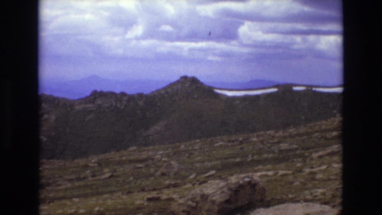 Rocky mountain ridge beneath a cloudy sky with snow patches in the distance.