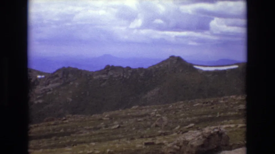 1973 Colorado 8mm frame with a rocky alpine slope, dark ridge line, distant snow, and layered clouds over the mountains.