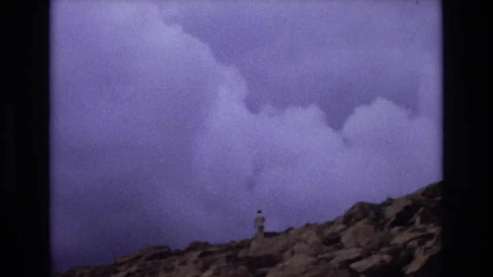 1973 Colorado 8mm frame showing a small hiker standing on rocky terrain beneath a dramatic cloudy sky in the mountains.
