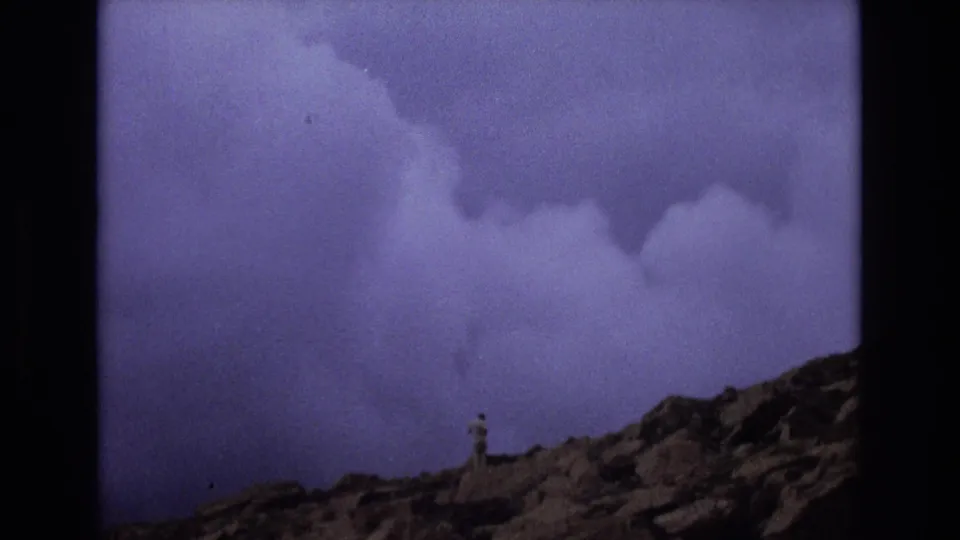1973 Colorado 8mm frame with a distant lone hiker on rocky mountain terrain under a large, dark cloud bank.