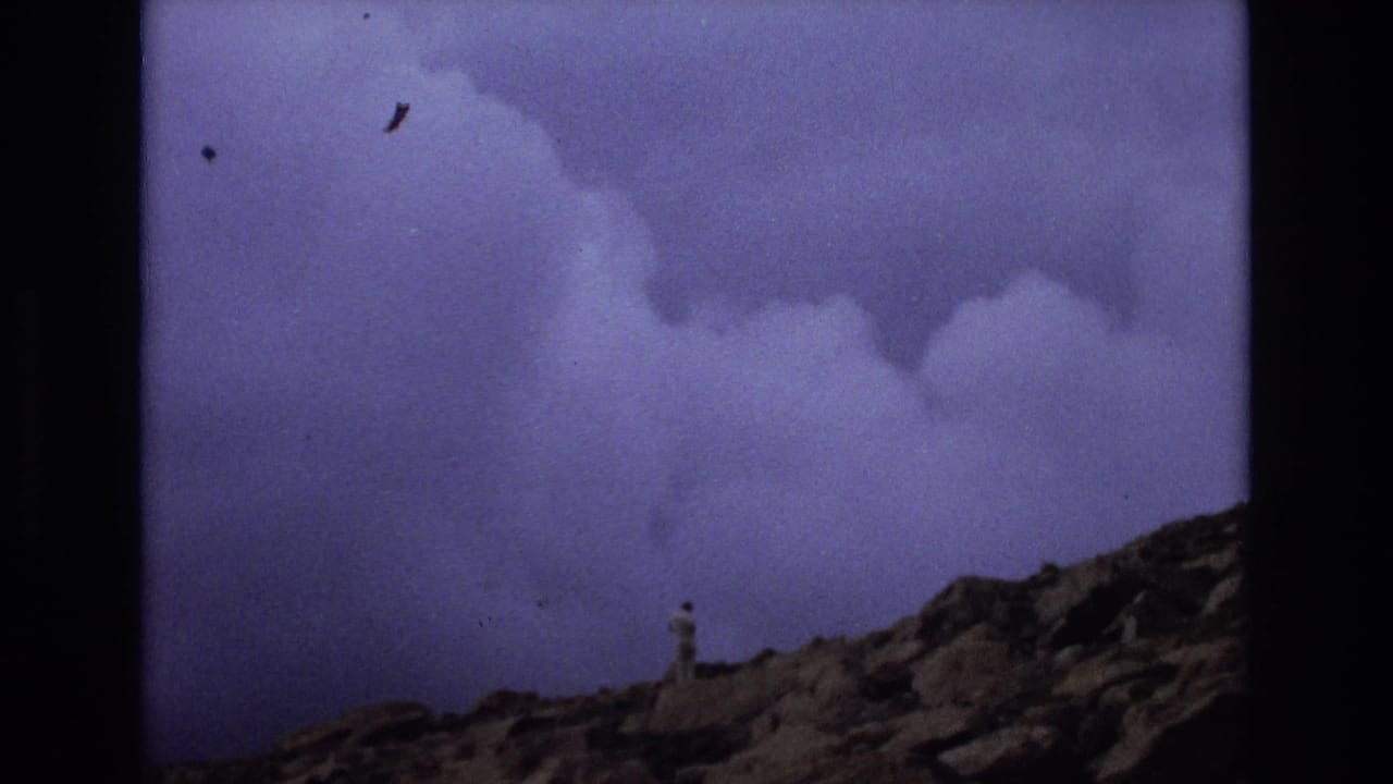 1973 Colorado 8mm frame showing a distant solitary man on rocky high ground under a heavy purple-blue sky, with dark film edges and visible grain.