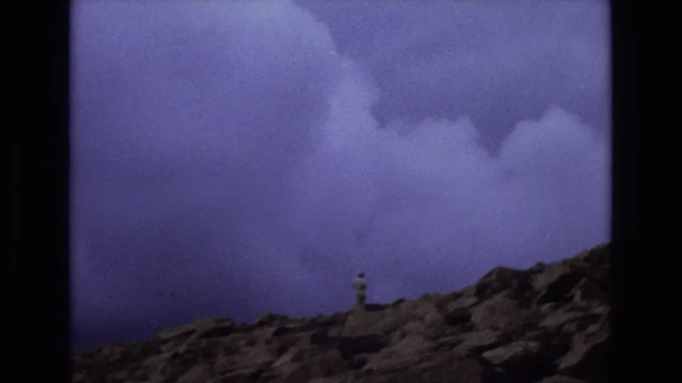 1973 Colorado 8mm frame with a distant man in light clothing standing on rocks while a stormy purple-blue cloud bank fills the background.