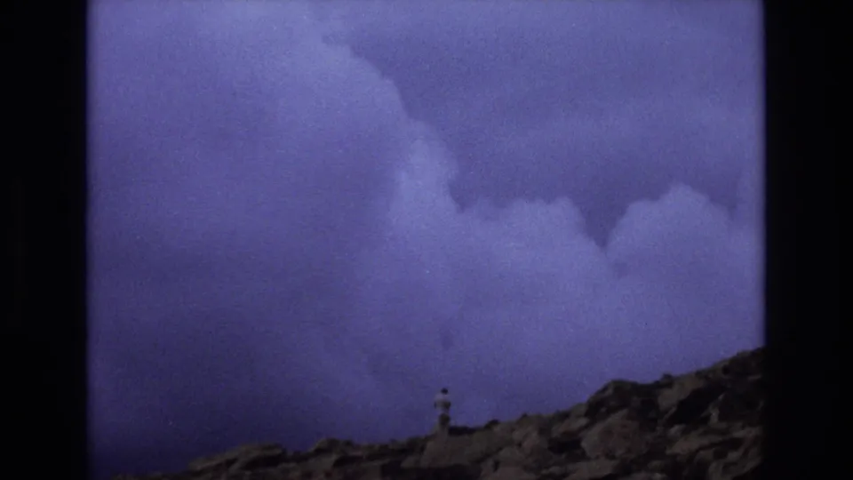 1973 Colorado 8mm frame showing a lone distant man near the lower edge of a rocky mountain scene, with dark purple cloud cover above.