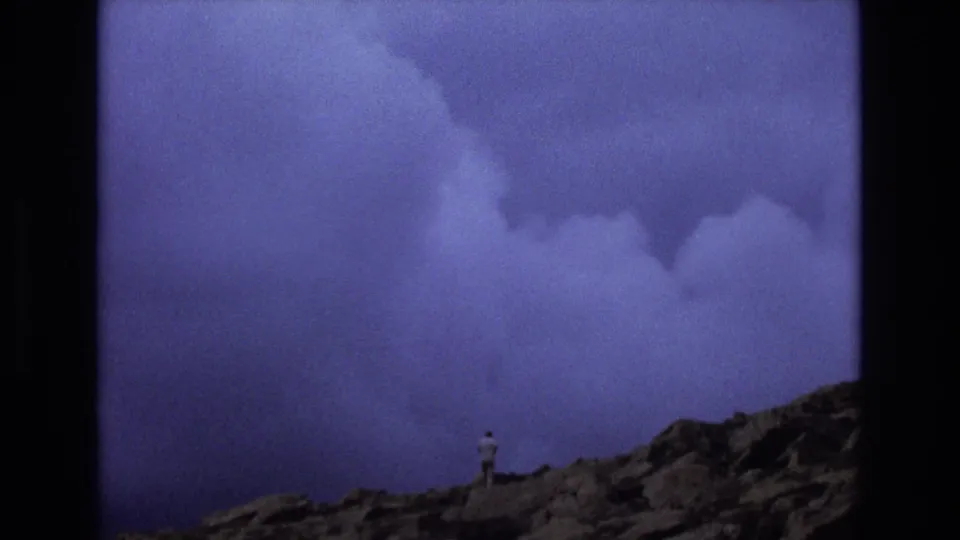 1973 Colorado 8mm frame with a small solitary figure on rocky high ground beneath an expansive purple-blue sky and heavy cloud forms.