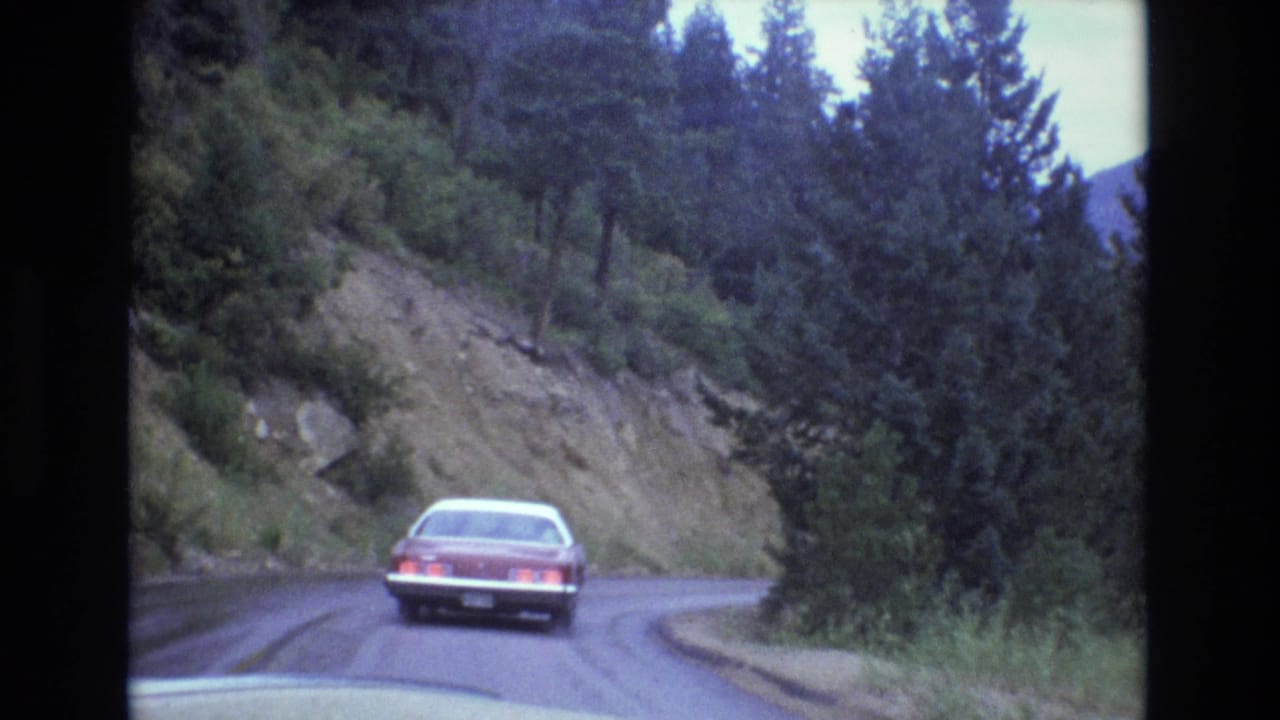 A red sedan drives away along a rain-darkened mountain road in Colorado, with evergreen trees, a rocky roadside slope, and soft 8mm film texture.