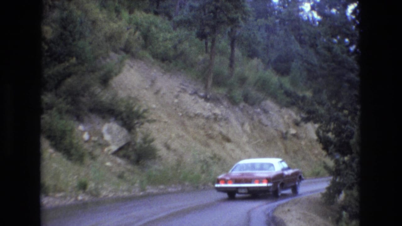Red sedan rounding a wet bend beside a rocky hillside and dense roadside trees.