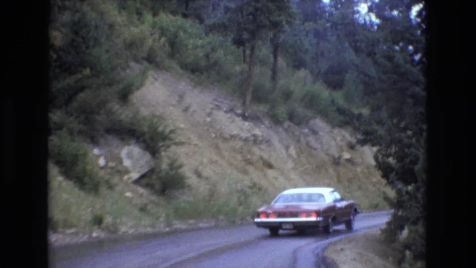 The red sedan rounds a damp mountain-road bend in Colorado, passing a steep rocky cut with trees and brush under cool overcast light.