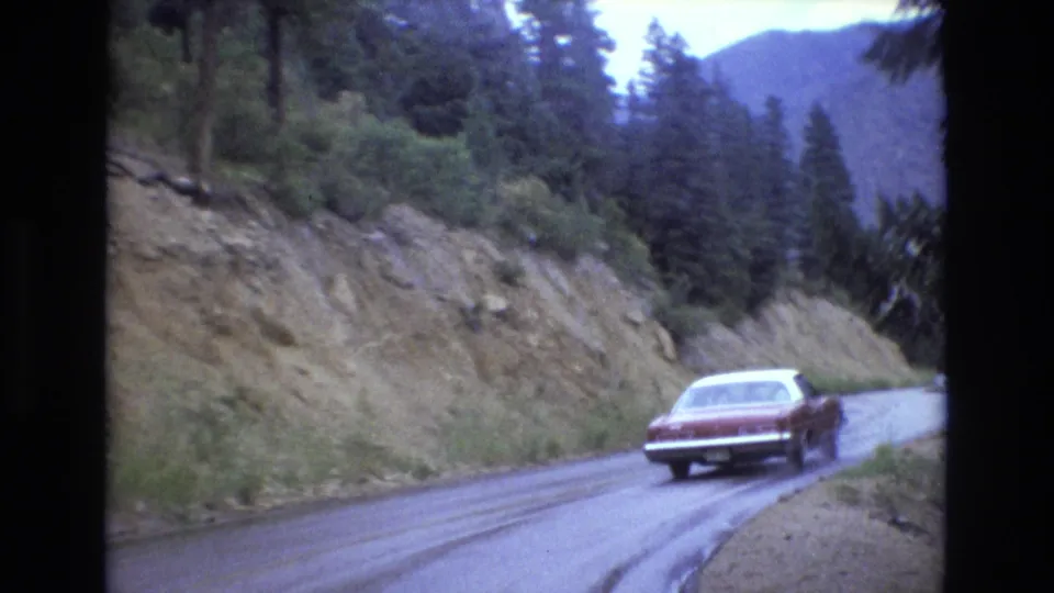 A red 1970s-era sedan continues along a slick mountain road in Colorado, framed by a rocky hillside, roadside vegetation, and evergreen trees.