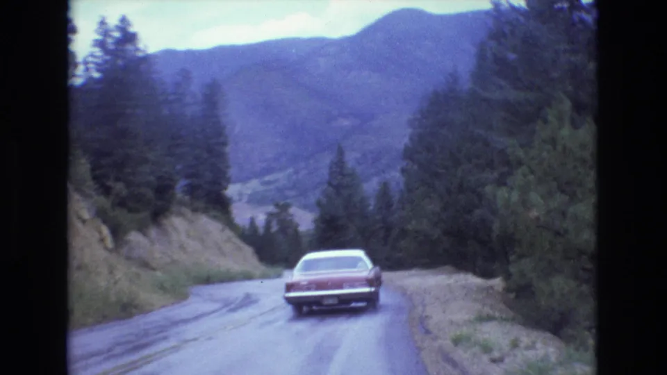 The red sedan drives away on a wet Colorado mountain road, with forested slopes and hazy distant mountains visible through the cool-toned 8mm frame.