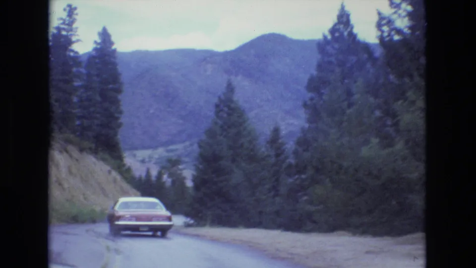 A red 1970s-era sedan descends a wet Colorado road toward a forested bend, with mountains and a valley visible through the trees.